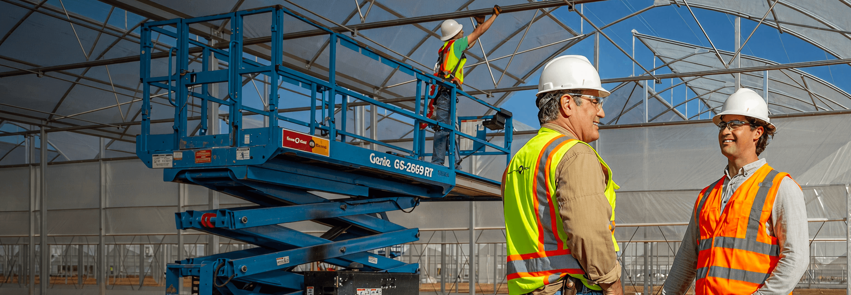 Two people talking on job site, with man on scissor lift in background.